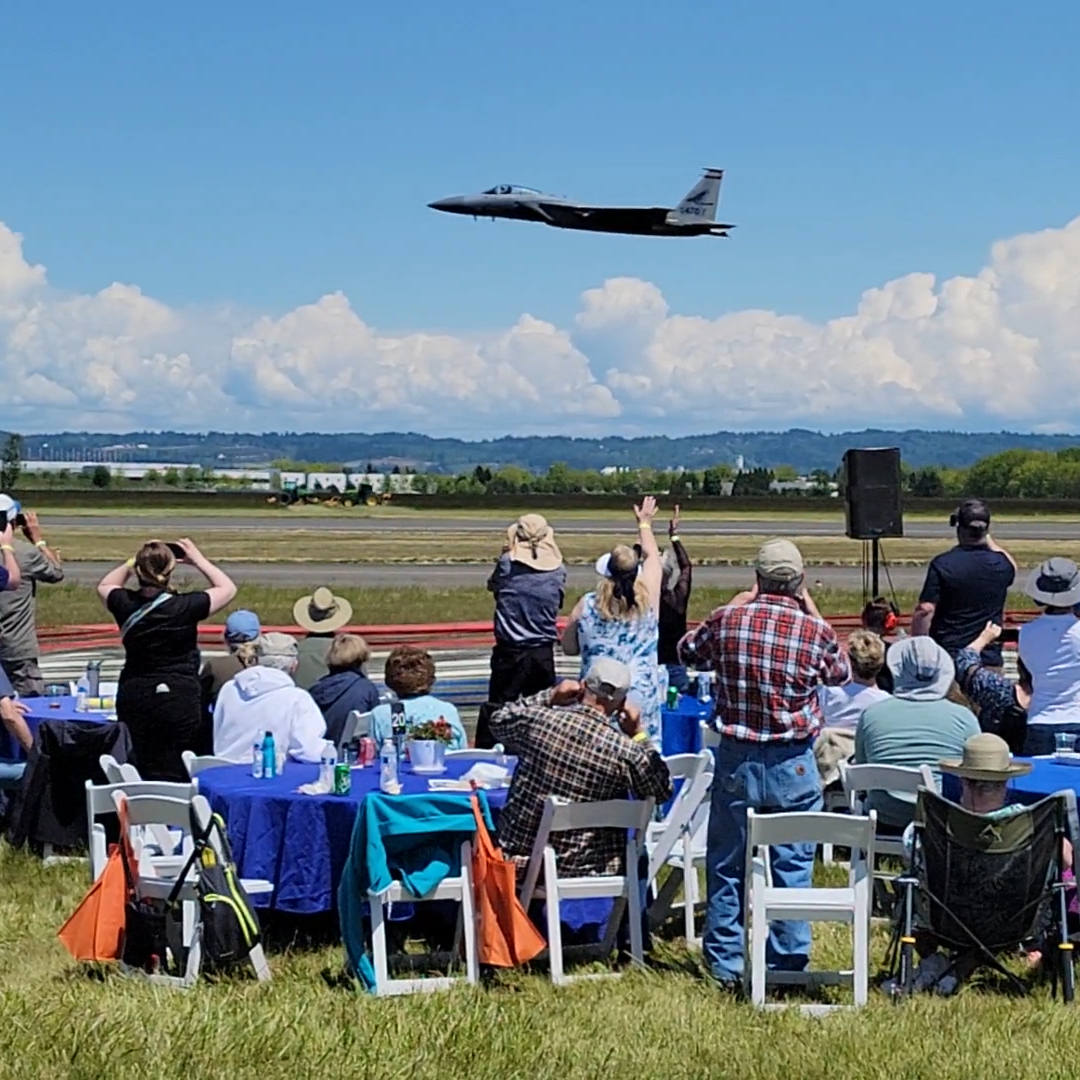 Oregon International Air Show - Ticket Type President's Club. Participants standing and seated next to tables on the flight line watching a military jet pass just in front of them in spectacular view.