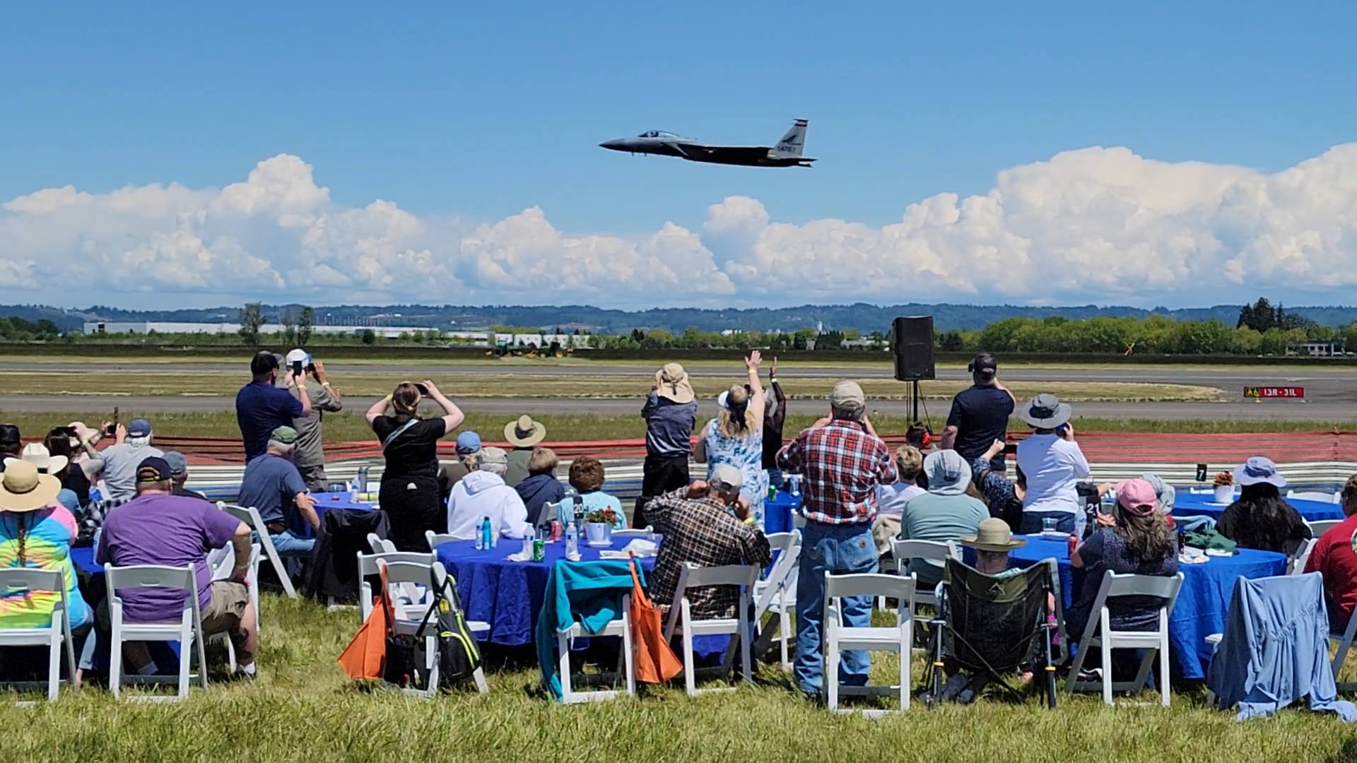 Oregon International Air Show - Ticket Type President's Club. Participants standing and seated next to tables on the flight line watching a military jet pass just in front of them in spectacular view.