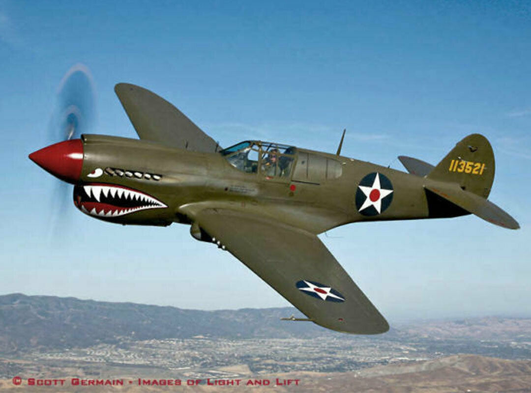 A WWII-era Curtiss P-40 Warhawk with signature shark-mouth nose art flies above the hills. The olive-drab warbird with U.S. roundels and red spinner pays tribute to the Flying Tigers and vintage American military aviation.