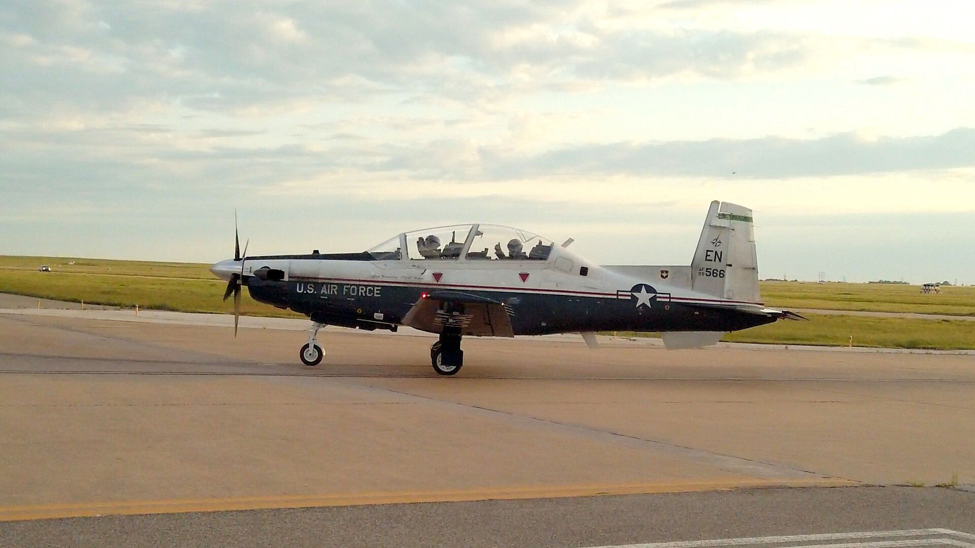 U.S. Air Force T-6 Texan trainer aircraft taxiing on a runway, with two pilots visible in the cockpit, painted in a black-and-white training livery at an airfield.