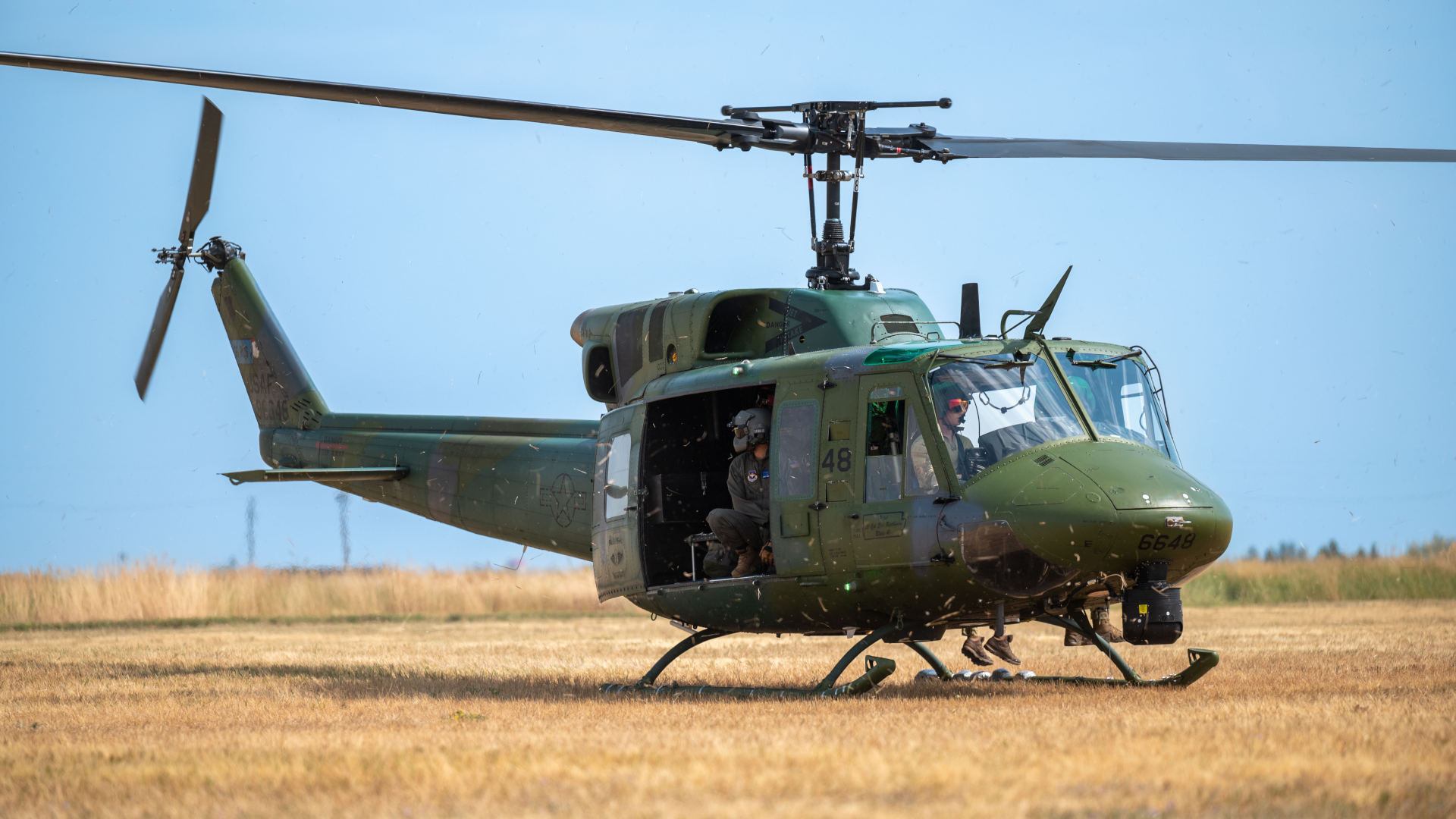 U.S. Air Force Bell UH-1 Huey helicopter hovering just above a grassy field, with crew members visible inside the open side door during a low-altitude operation.