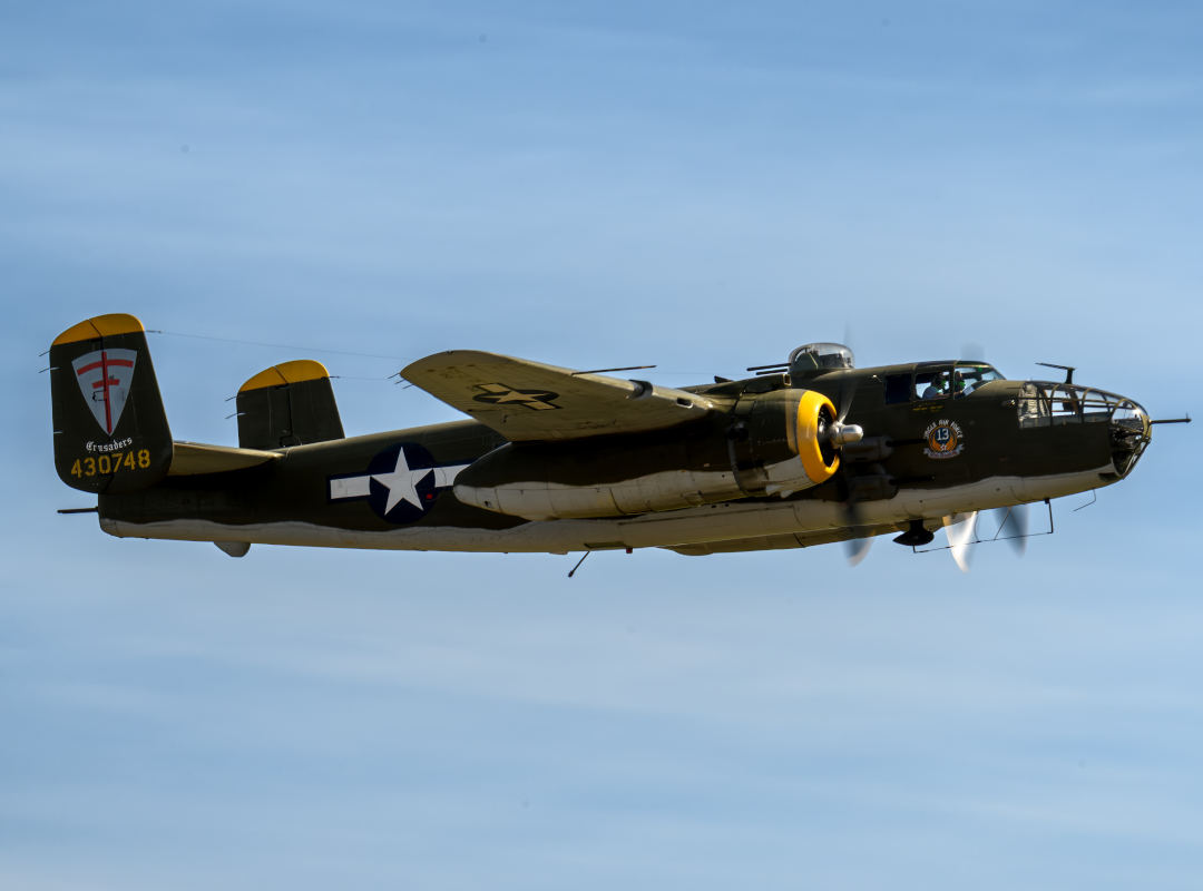 B-25 Mitchell twin-engine bomber flying in clear blue sky, showing olive drab fuselage, U.S. star insignia, and spinning propellers in side profile