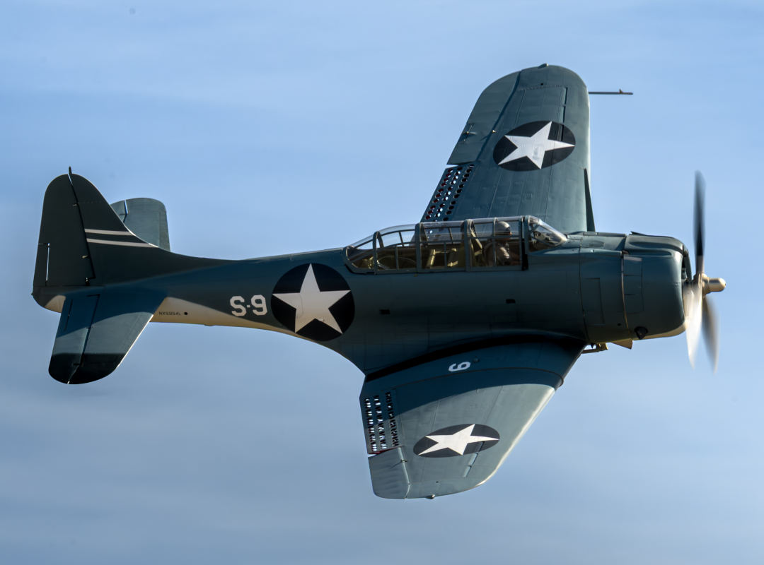 SBD Dauntless dive bomber from the Erickson Aircraft Collection flying in side profile against a clear blue sky, showing dark green fuselage, U.S. star insignia, glass canopy, and spinning propeller