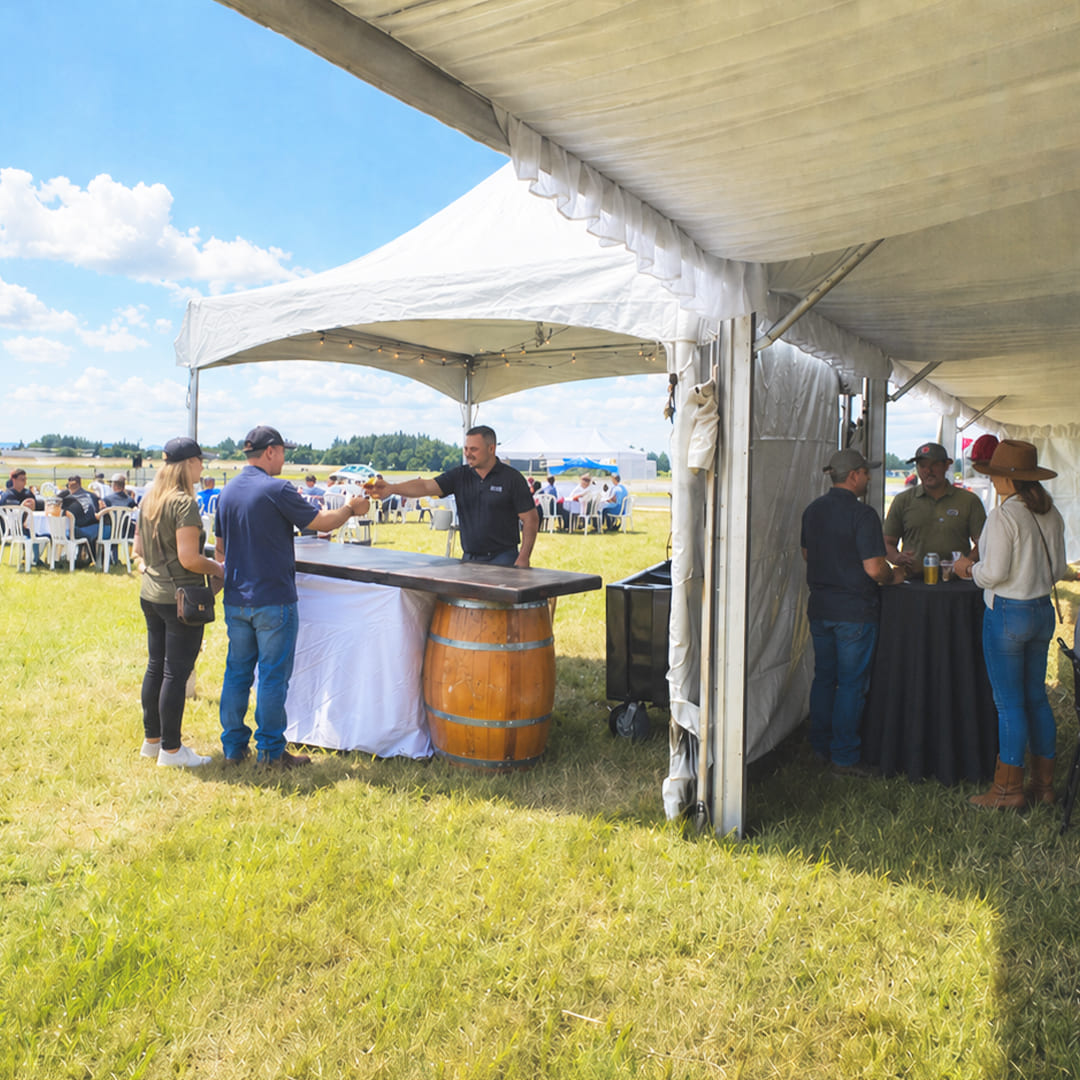 Oregon International Air Show - Ticket Type Presidents Club. Looking out from inside the shade tent to the seating area on the flight line. A couple stands at a bar getting a drink from a bartender. Some people stand around a cocktail table talking inside the tent.