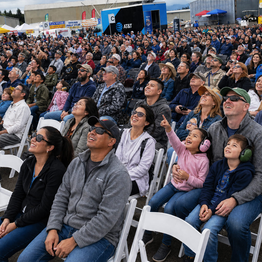 Oregon International Air Show - Ticket Type Reserved Seating. A large gathering of people all sit in rows of chairs, pointed in the same direction and excitedly looking toward the sky. Air show grounds are seen off in the distance. 