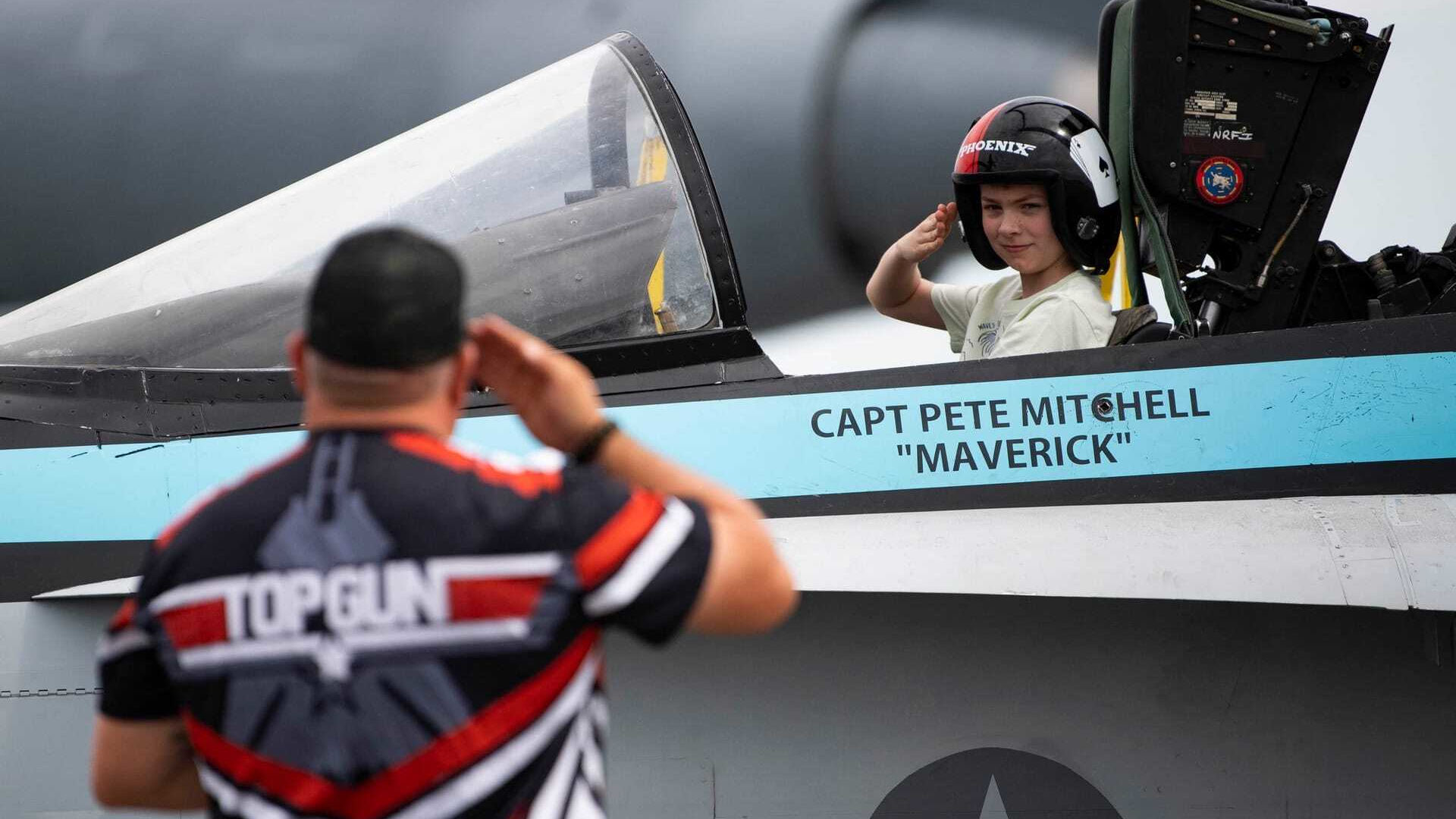 Child wearing a flight helmet sits in a mock F/A-18 cockpit labeled “Capt Pete Mitchell ‘Maverick’” during the Top Gun cockpit experience by DreamBig Entertainment, saluting an adult in a Top Gun shirt who returns the salute, with aircraft canopy and controls visible in the foreground