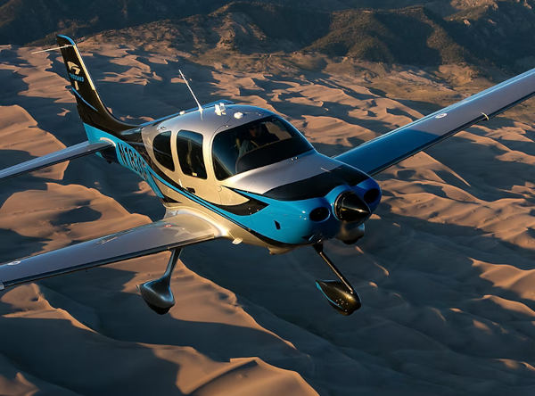 Blue and white Cirrus SR series aircraft flying above desert sand dunes with mountains in the distance under warm light.