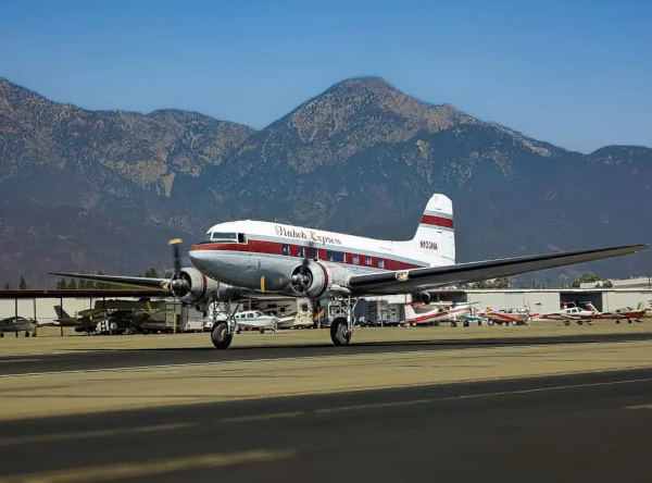 Vintage Douglas DC-3 aircraft in red and white livery taxiing on a runway with mountains and parked planes in the background.
