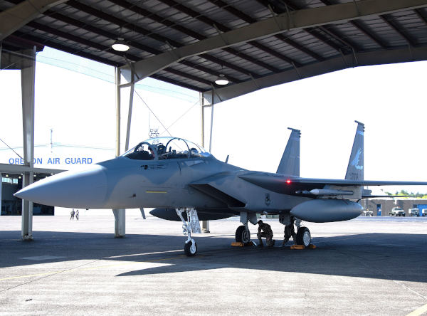 Gray F-15 fighter jet parked under a large hangar with ground crew beneath the aircraft and Oregon Air Guard signage visible in the background.