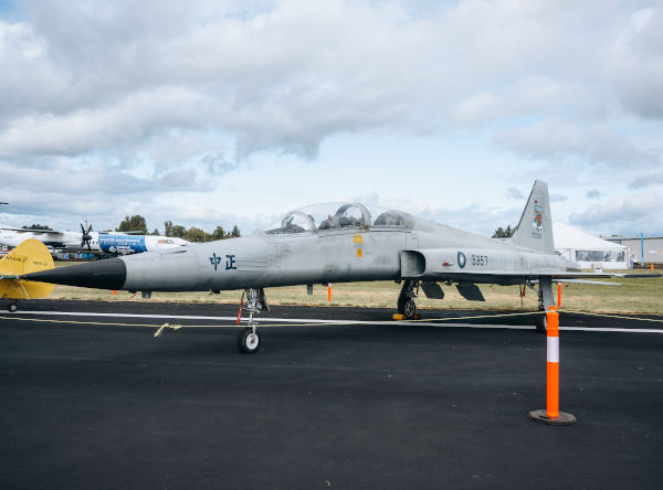 Gray Northrop F-5 fighter jet parked on an airfield ramp with landing gear down and orange safety cones nearby, featuring Chinese characters on the fuselage and a cloudy sky overhead.