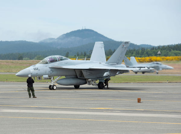 Gray EA-18G Growler taxiing on a runway with ground crew nearby and another jet blurred in the background.