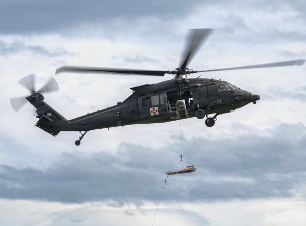 HH-60 Black Hawk helicopter hovering in cloudy sky while crew member operates hoist lowering equipment below the aircraft.