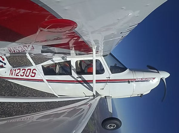 American Champion Super Decathlon N123DS captured from wing-mounted perspective in flight with red and white livery against a clear blue sky.
