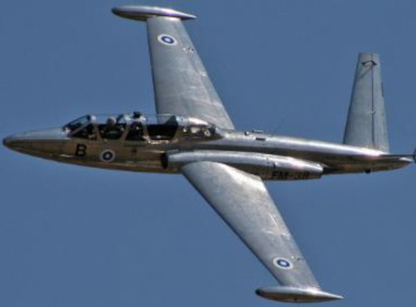 Fouga CM-170 Magister jet in flight, silver with French roundels, distinctive V-tail design, viewed from below against a clear blue sky.