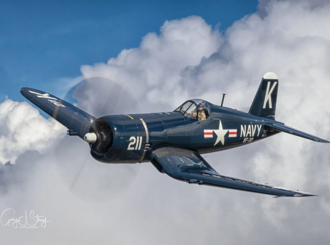 F4U Corsair warbird in dark U.S. Navy markings flies past towering white clouds, with the pilot visible in the cockpit and the propeller blurred in motion.
