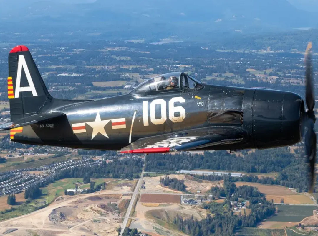 F8F Bearcat warbird in dark Navy markings flies above a rural landscape, with the pilot visible in the cockpit and the propeller spinning.