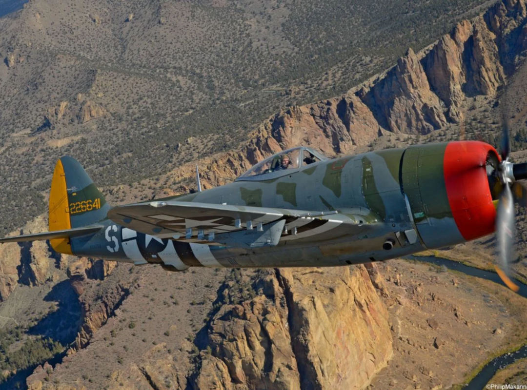 P-47 Thunderbolt warbird in camouflage paint flies along a rocky canyon landscape, with the pilot visible in the cockpit and the propeller blurred in motion.