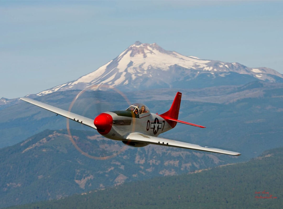 P-51 Mustang flying over forested mountains with a snow-capped peak in the background.