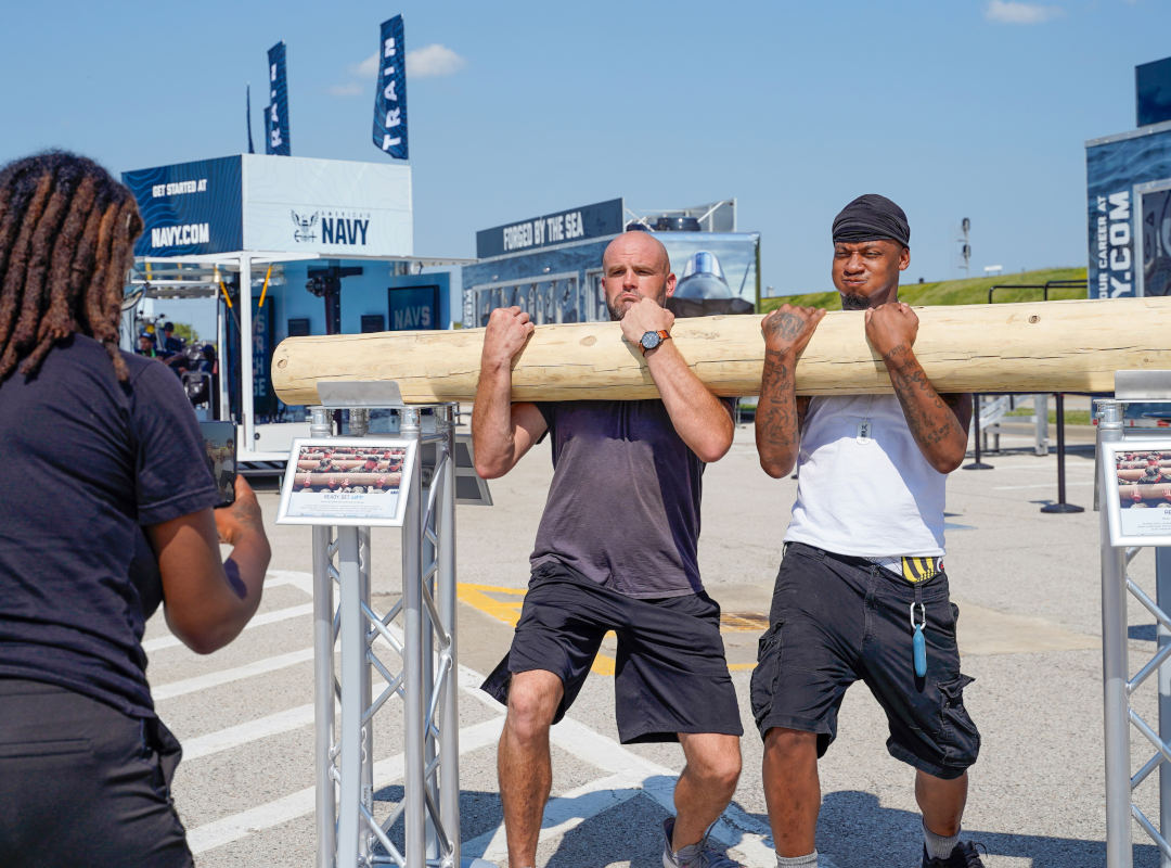 Two participants lift a large wooden training log while a Navy representative observes during a physical challenge.