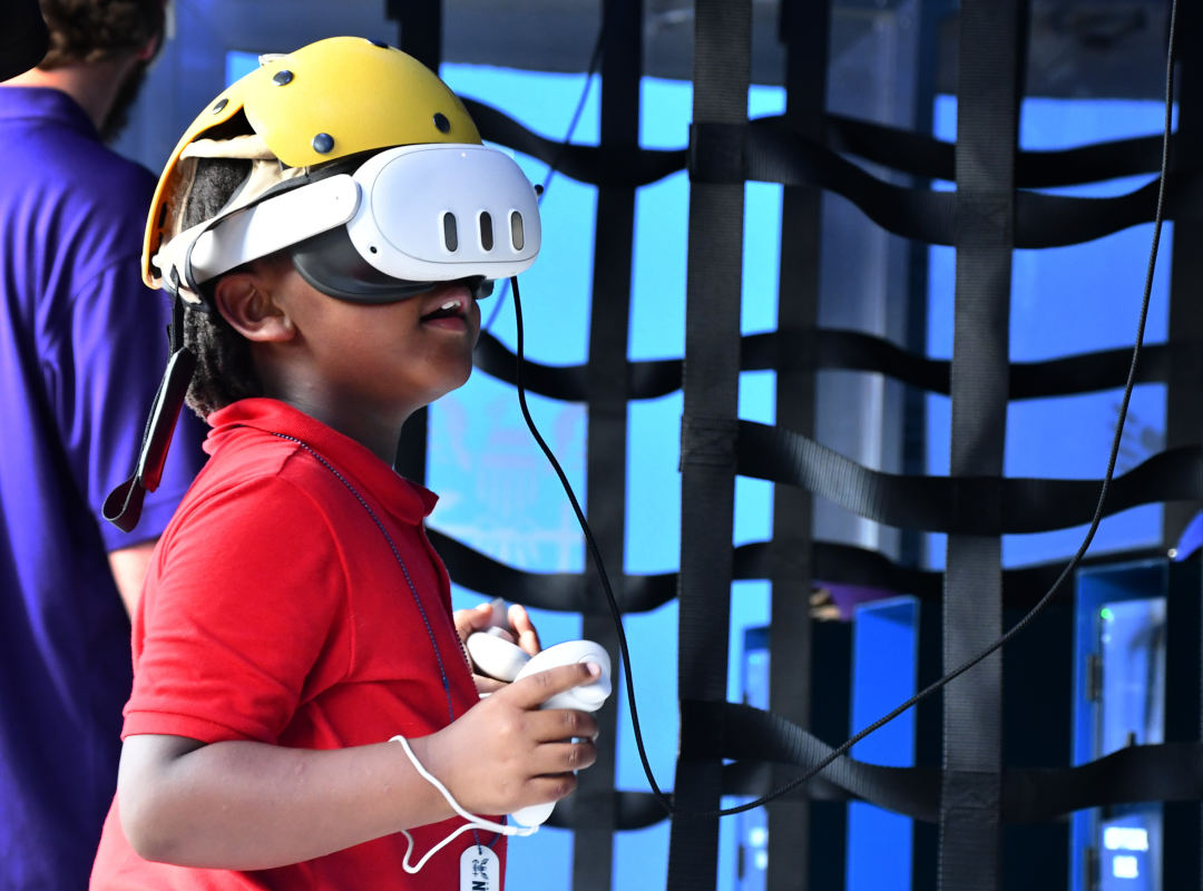 A child wearing a virtual reality headset and protective helmet holds two controllers inside the U.S. Navy interactive experience.